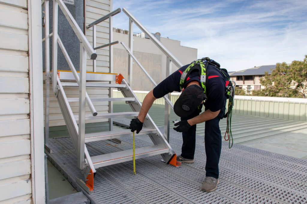 Compliance technician measuring the height from the landing to the first step on a staircase using a tape measure.