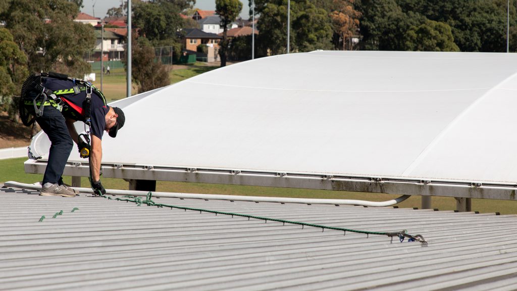 Worker using a roof anchor system at a sports facility in Sydney.
